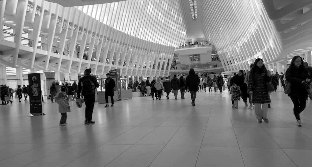 The interior of the Oculus at the World Trade Center in New York City, showcasing its sleek white design and soaring, rib-like structure designed by Santiago Calatrava. The vast, open space is filled with people walking, commuting, and shopping within the Westfield World Trade Center mall. As a major transit hub, the Oculus connects PATH train passengers and subway riders while serving as an architectural landmark in Lower Manhattan. The bright, modern aesthetic creates a striking contrast to the bustling movement of daily life.